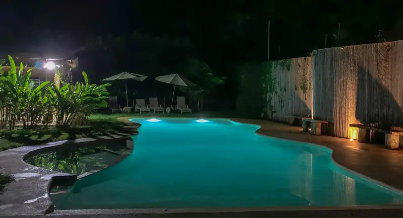 Piscina iluminada de noche entre plantas, con sombrillas y reposeras