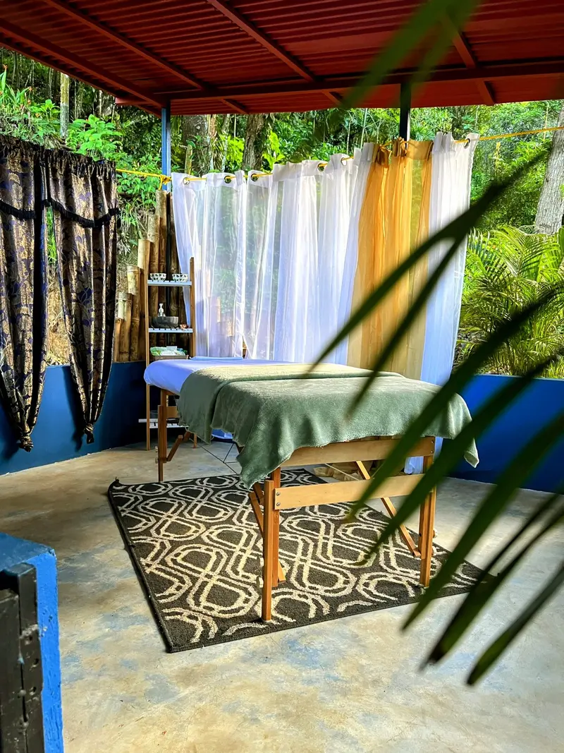 Estudio de masajes al aire libre, con camilla cubierta de tela verde, cortinas blancas y una alfombra estampada