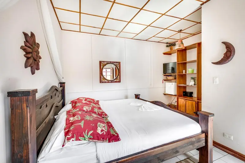 Habitación con una cama de madera con colcha roja de flores, un espejo y estantes en la pared