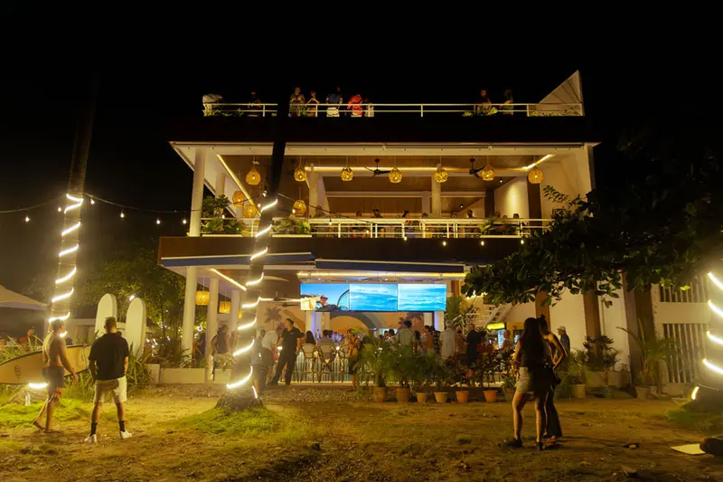 Fachada de Ozeano iluminada de noche, con tablas de surf y luces en las palmeras