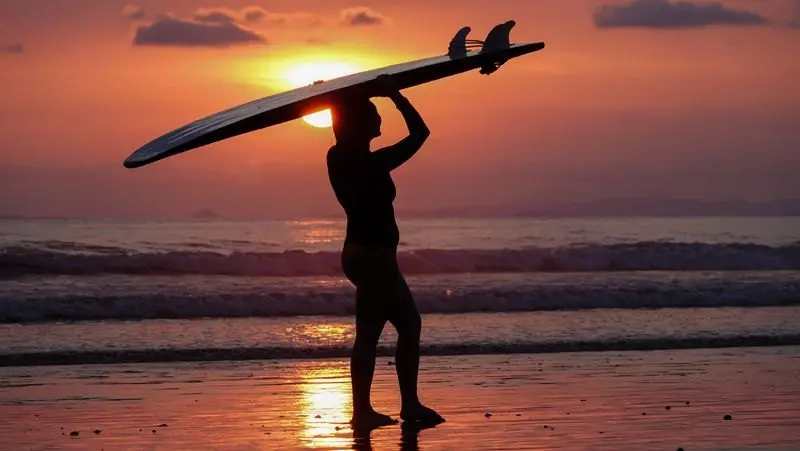 Silueta de una persona con la tabla sobre la cabeza frente a un atardecer rojo