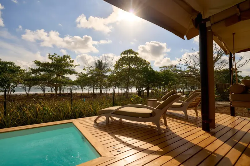 Terraza de madera con una reposera junto a una piscina privada turquesa y la playa al fondo