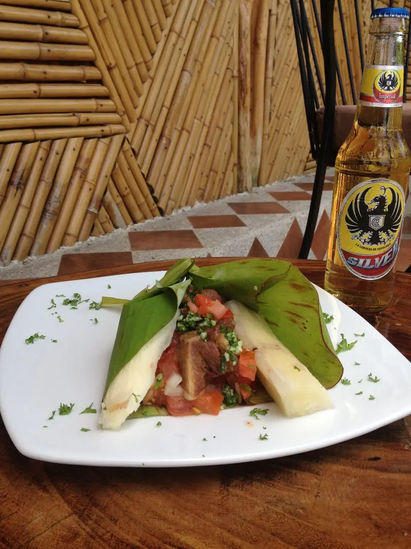 Plato blanco con un bocado servido sobre hoja de banano con tomate picado, junto a una botella de cerveza