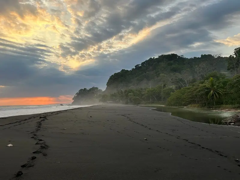Playa de arena oscura al amanecer con nubes de colores y la costa verde al fondo