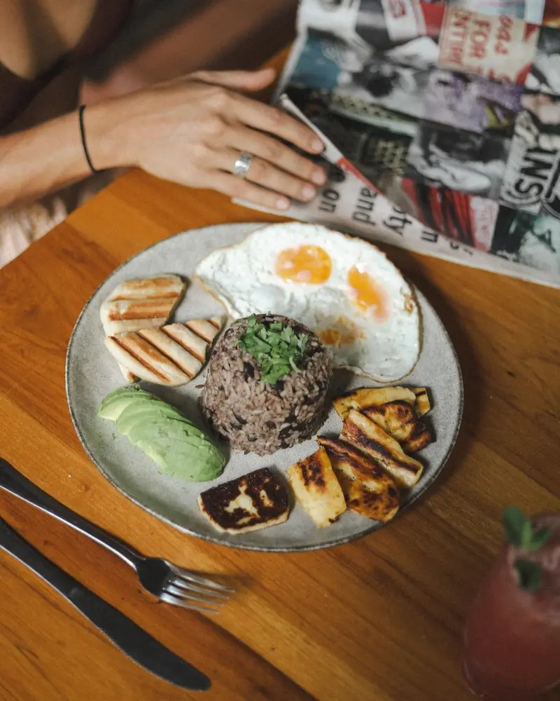 Plato de desayuno con huevos fritos, gallo pinto, aguacate y queso frito junto a una revista