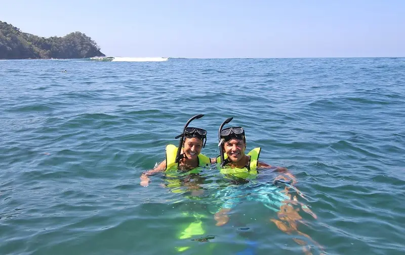 Dos personas con máscara de snorkel y chaleco amarillo en el mar con la costa al fondo