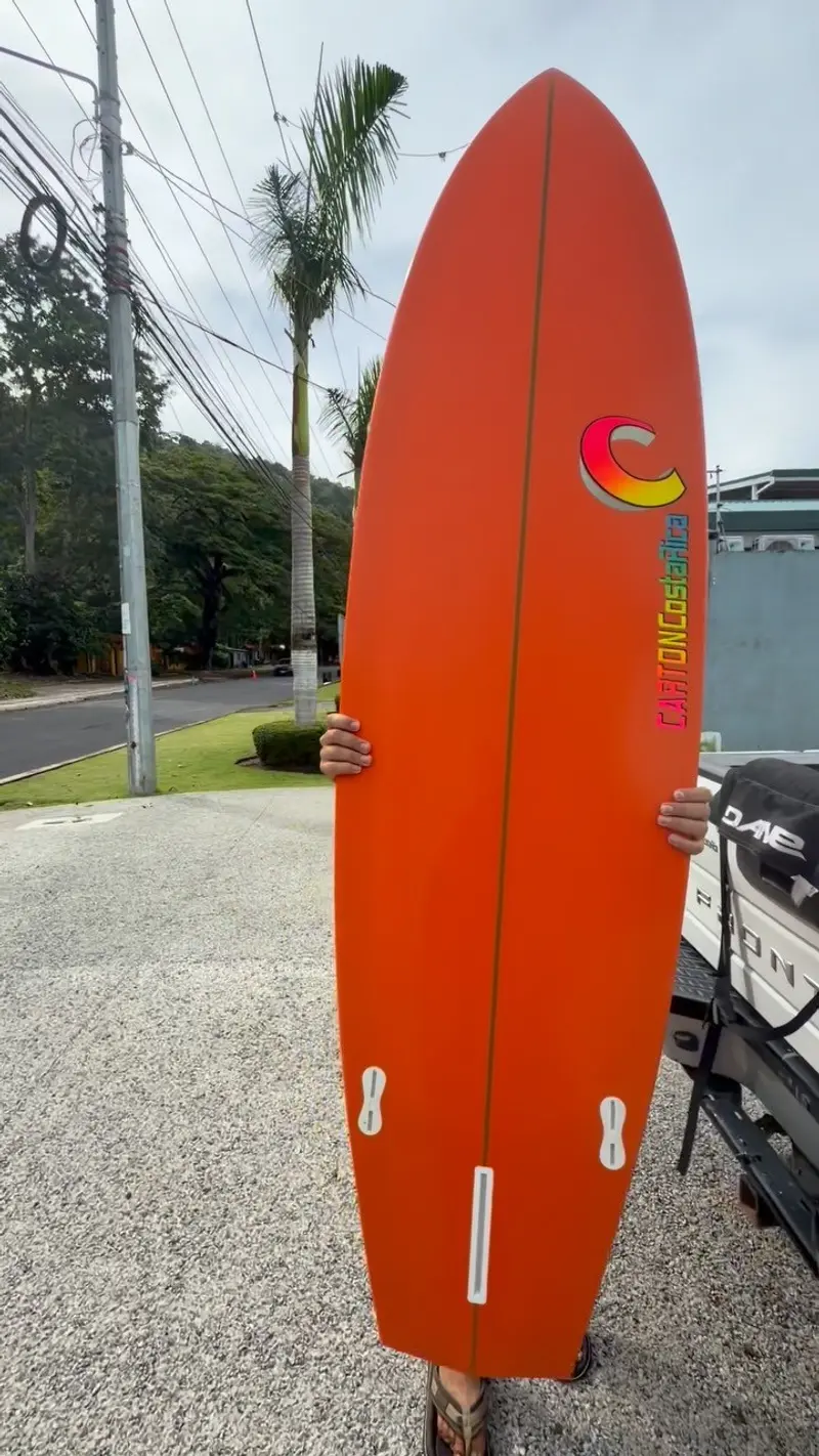 Tabla de surf naranja sostenida en vertical frente al taller, con el logo de Carton y aletas visibles