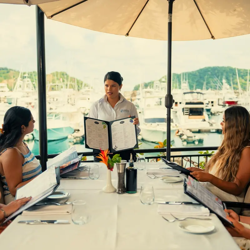 Mesera mostrando el menú a comensales en la terraza frente a la marina
