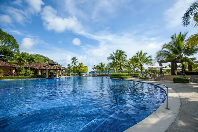Piscina grande de borde curvo con palmeras y techos de palapa bajo un cielo con nubes