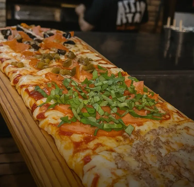 Pizza por metro con albahaca fresca y tomate sobre una tabla larga, con un cocinero al fondo