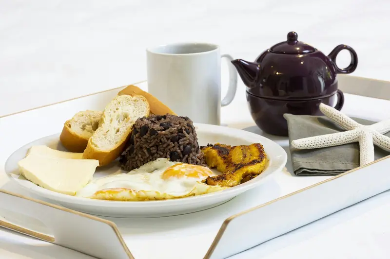 Desayuno servido en una bandeja con huevos, queso, pan, gallo pinto y plátano maduro, y una taza de café