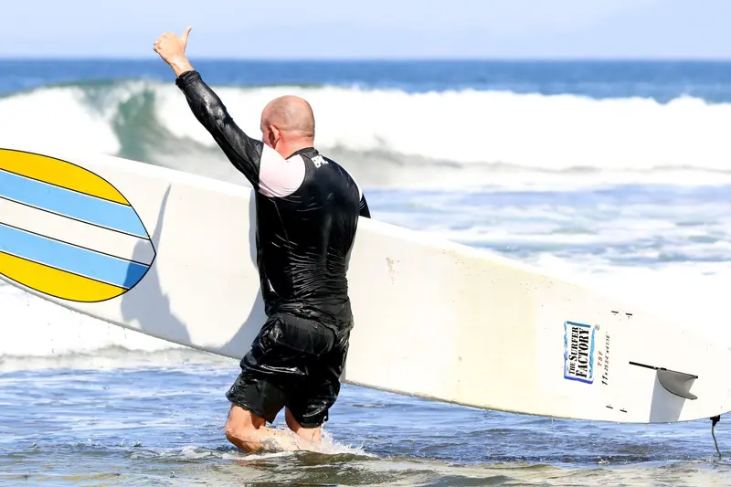 Surfista con licra y tabla blanca entrando al mar con un brazo en alto, con una ola de fondo