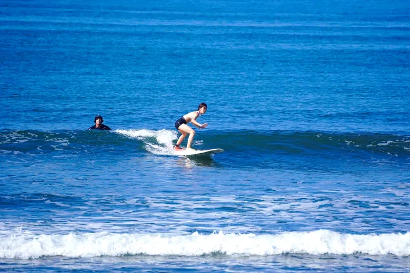 Surfista de pie sobre una ola azul en Jacó, con otra persona esperando en el agua