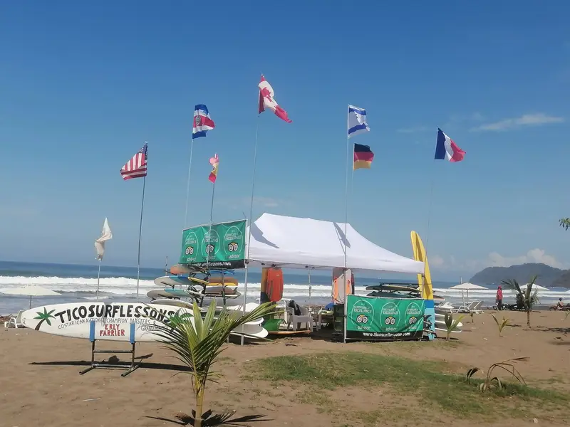 Carpa blanca con tablas de surf alineadas en la arena y banderas de varios países junto al mar
