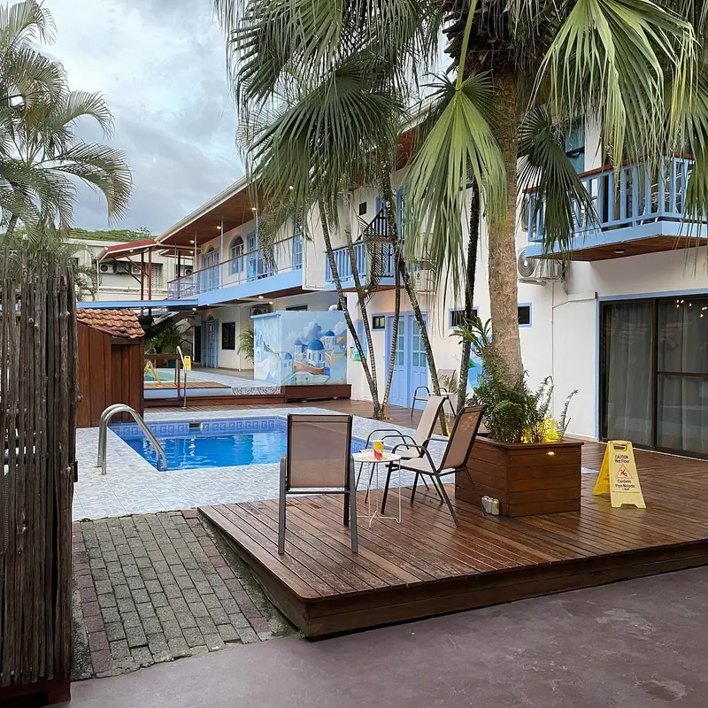 Patio con piscina y terraza de madera entre palmeras, con los balcones de las habitaciones al fondo