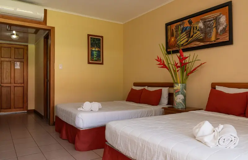 Habitación con dos camas con cojines rojos, aire acondicionado, un cuadro en la pared y la puerta de entrada