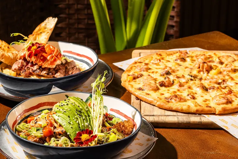 Dos bowls con ensalada, carne y aguacate junto a una pizza sobre una mesa de madera