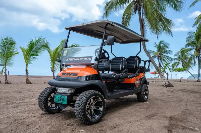 Carrito de golf negro y naranja de seis plazas sobre la playa, con palmeras detrás