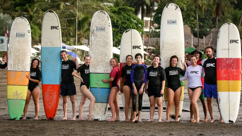 Grupo de alumnos con licras de surf posando junto a tablas de colores paradas en la arena