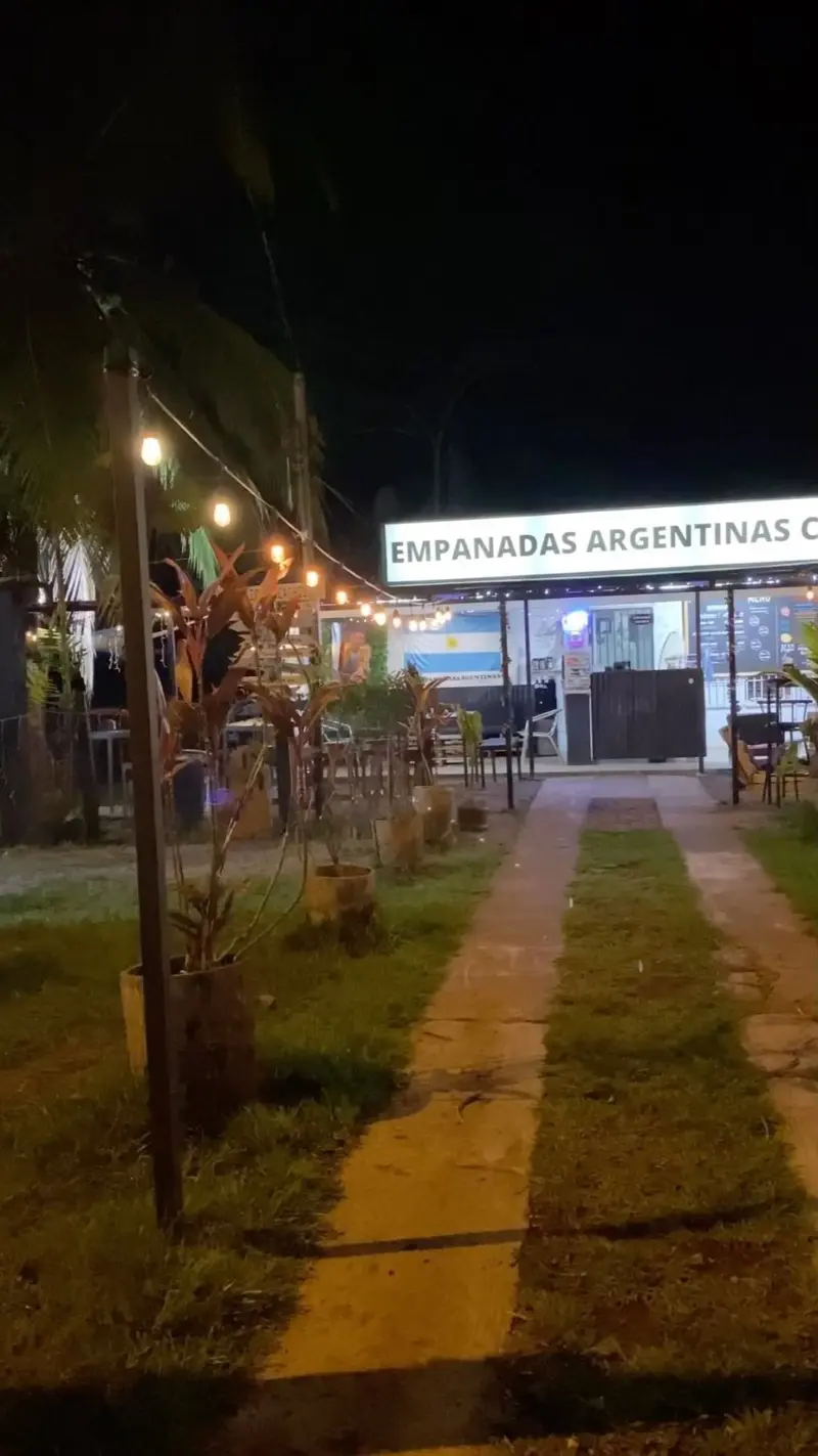 Entrada del local de noche, con un camino de tierra, luces colgantes y el cartel Empanadas Argentinas