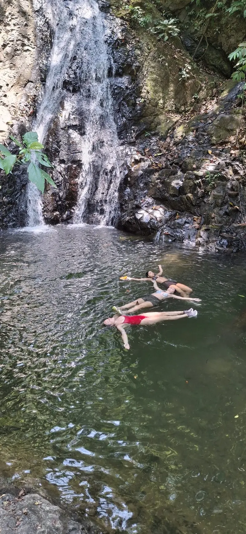 Tres personas flotando en una poza de agua verde al pie de una cascada
