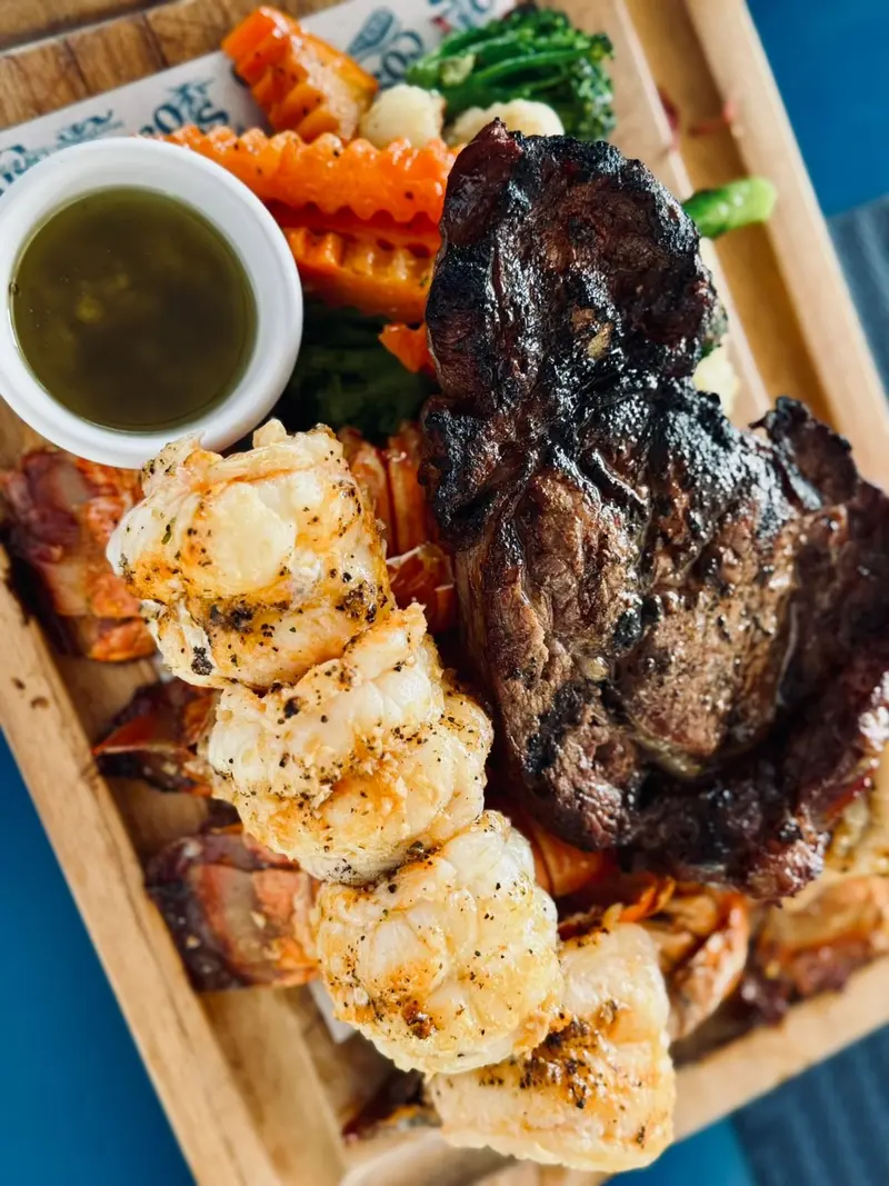 Tabla de madera con un corte de res a la parrilla, colas de langosta, vegetales y una salsa verde