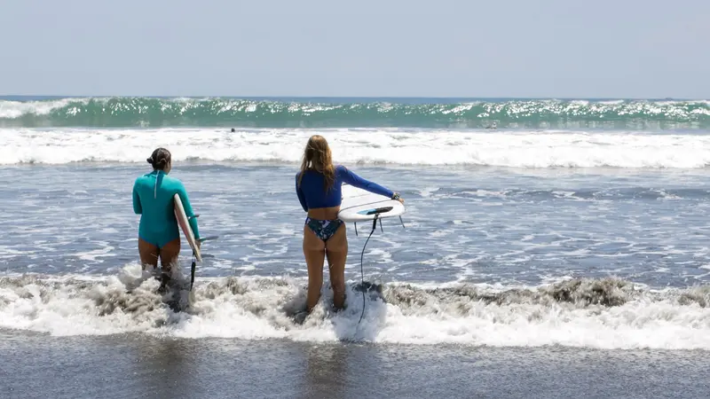 Dos personas con tablas de surf caminando por la orilla hacia el mar, vistas de espaldas