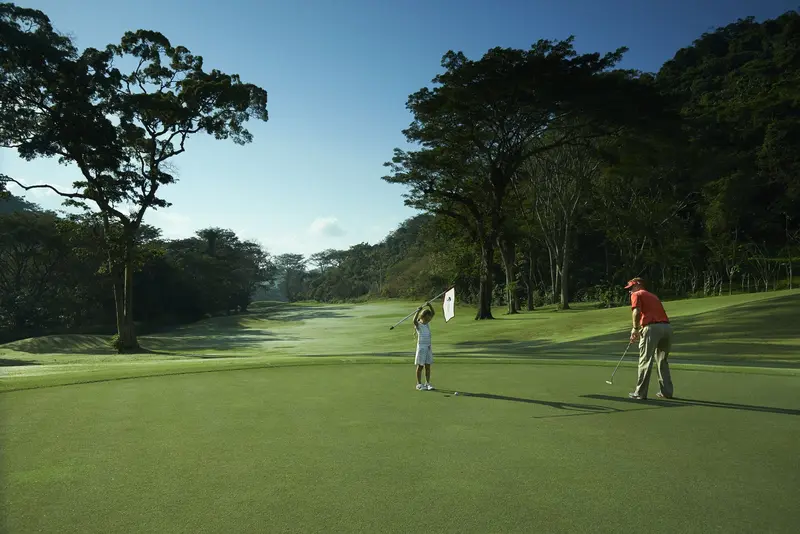 Dos golfistas en un green, uno de ellos con camisa roja, con el bosque al fondo