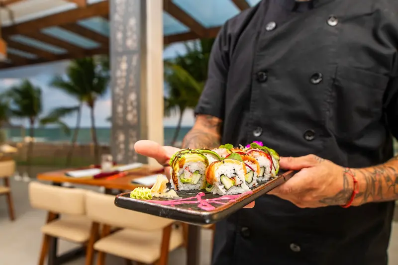 Cocinero con chaqueta negra sosteniendo una bandeja de rolls de sushi, con el océano al fondo