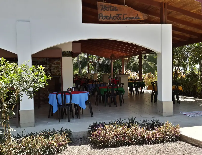 Restaurante al aire libre con mesas bajo un techo de madera y arcos blancos, entre jardines