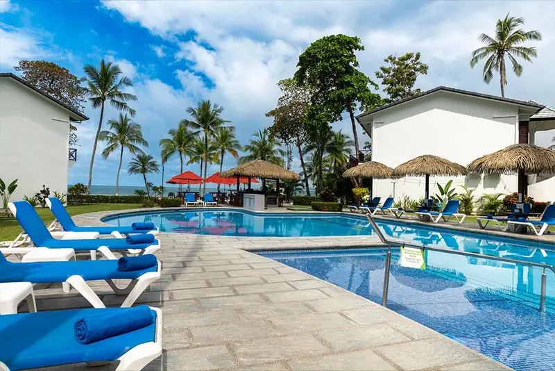 Piscina con tumbonas azules y sombrillas de palma, con los edificios del hotel entre las palmeras