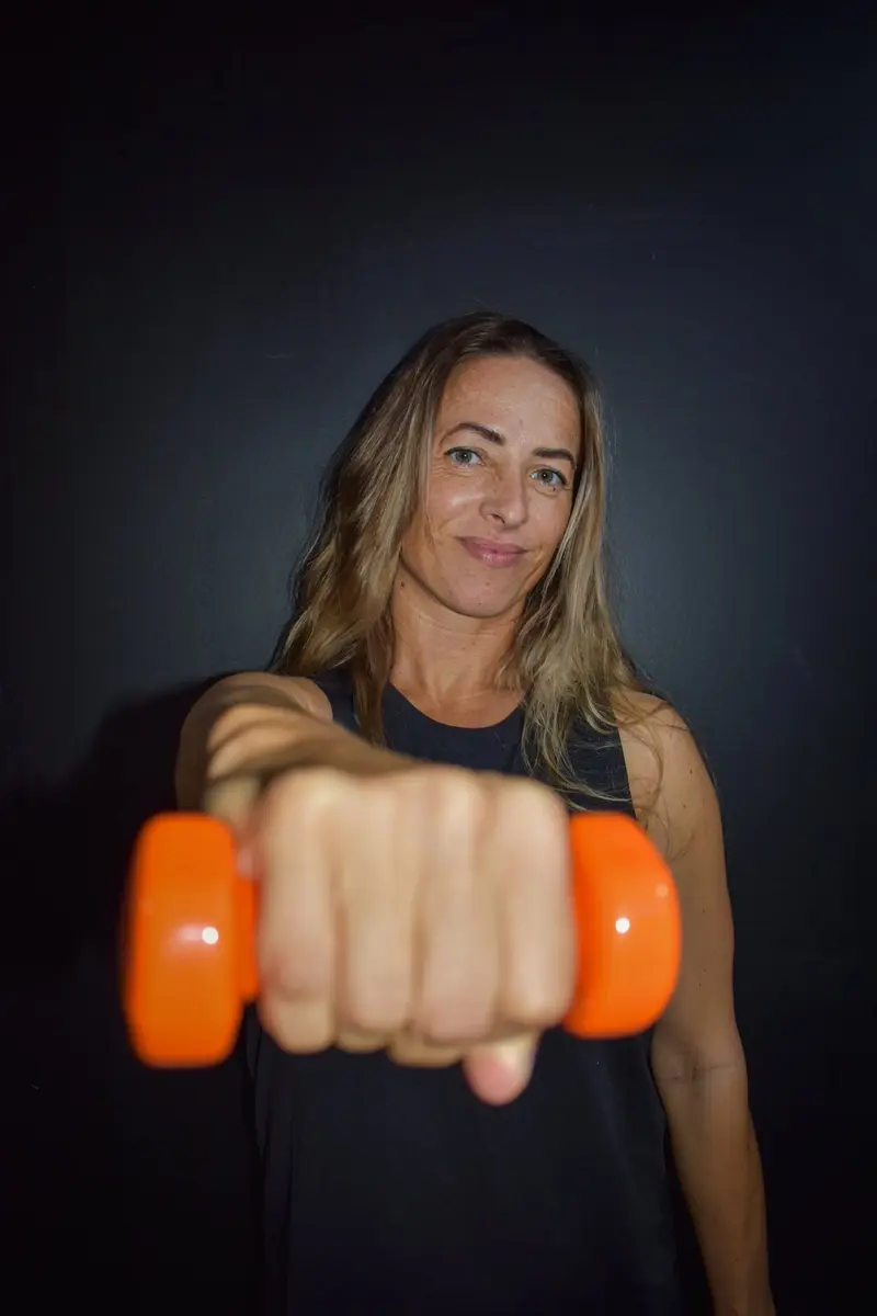 Instructora sonriendo con una mancuerna naranja extendida hacia la cámara, sobre fondo oscuro