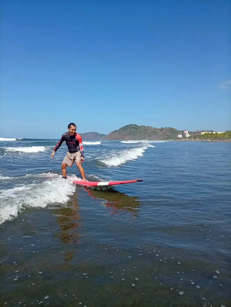 Surfista de pie sobre una tabla roja en una ola pequeña, con una colina al fondo