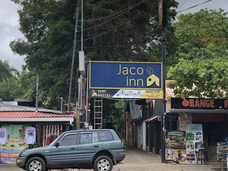 Rótulo azul de Jaco Inn Surf Hostel sobre la calle, con un auto estacionado y tiendas alrededor