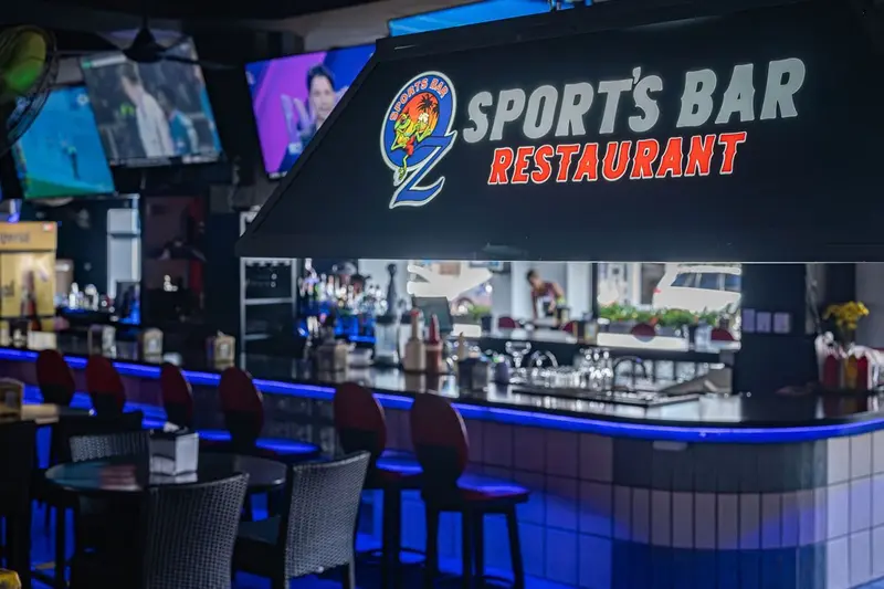 Barra del Oz Sports Bar con taburetes, luces azules y pantallas sobre el rótulo del restaurante