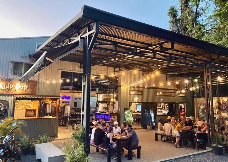Terraza techada de la cervecería con luces colgantes y mesas de madera con clientes