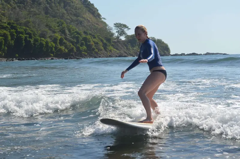 Mujer surfeando una ola pequeña con una tabla blanca frente a una costa con selva