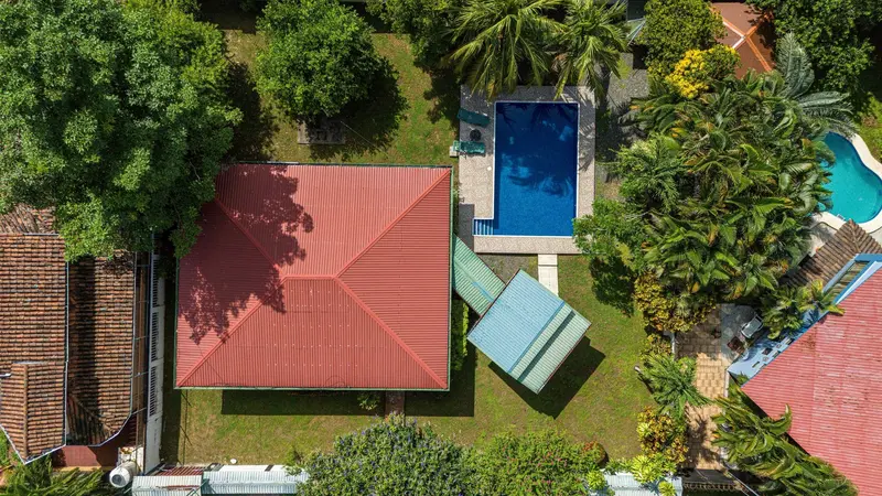 Vista aérea de una casita de techo rojo entre jardines, con una piscina azul y camastros al lado