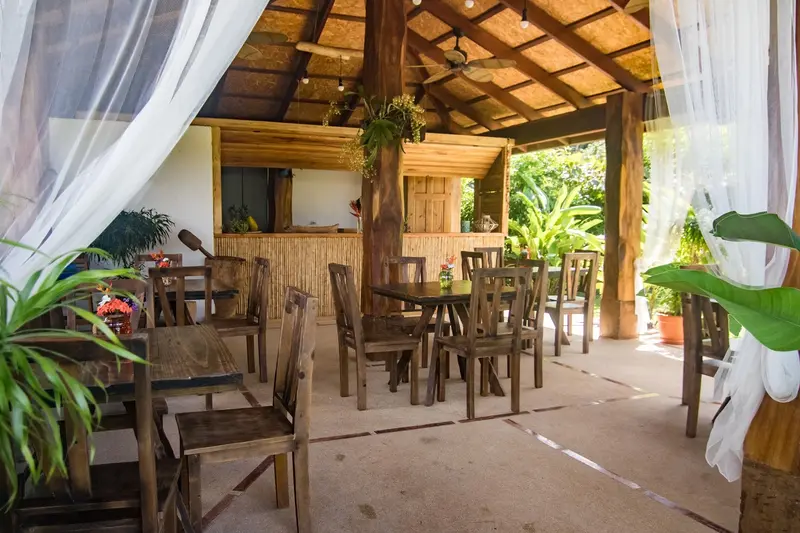Rancho comedor de techo de madera con mesas y sillas, cortinas blancas y jardín