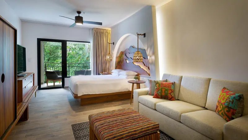 Habitación con cama, sofá con cojines de colores, televisor y puerta de vidrio al balcón