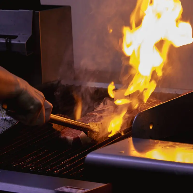 Cocinero moviendo la carne con unas pinzas sobre una parrilla con llamas