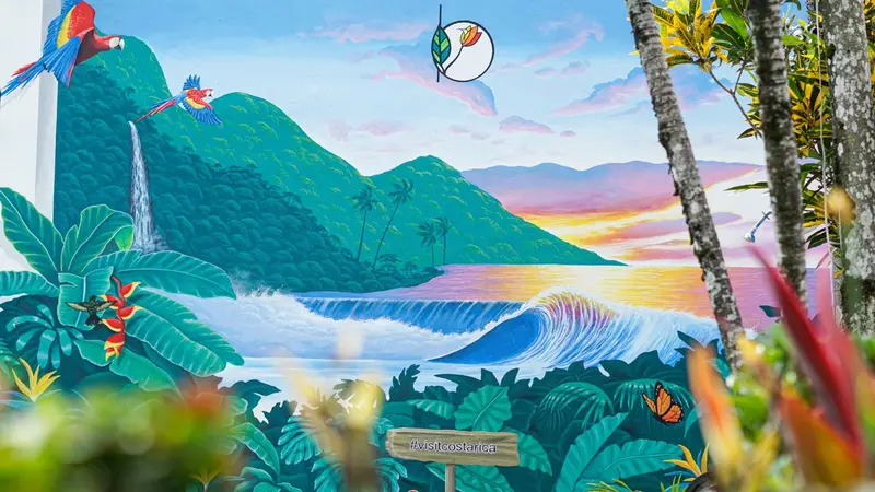 Mural de una ola frente a montañas verdes con guacamayas y atardecer rosado, pintado en la pared de la piscina
