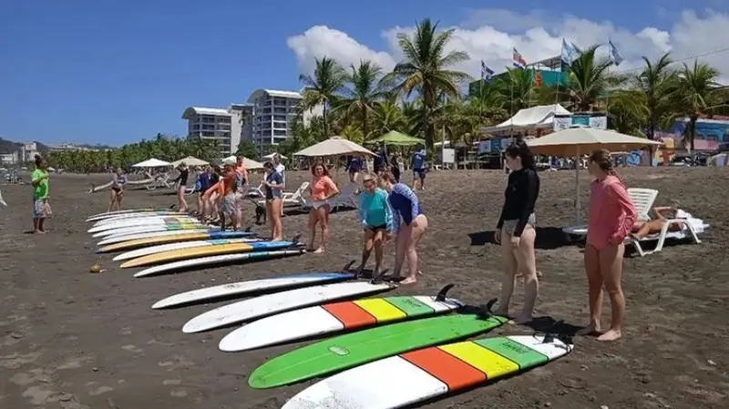 Tablas de surf de colores alineadas en la arena oscura de la playa, con bañistas y carpas al fondo