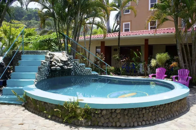 Piscina redonda con escalones azules y borde de piedra, rodeada de jardín y de las habitaciones