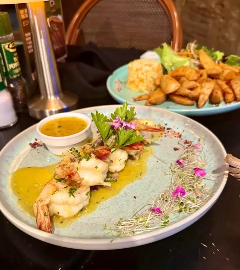 Camarones en salsa amarilla con germinados y flores sobre un plato celeste, con una fuente de papas y arroz al fondo