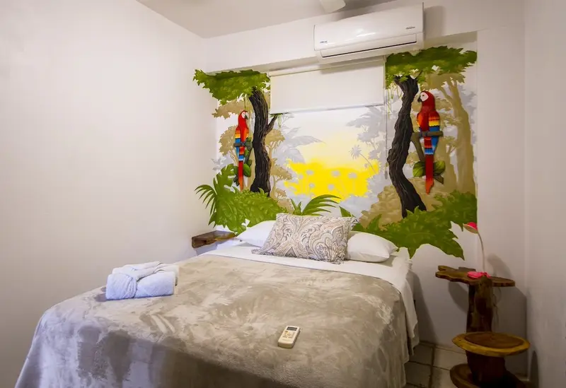 Habitación con cama, ventana y mural de guacamayas en la pared