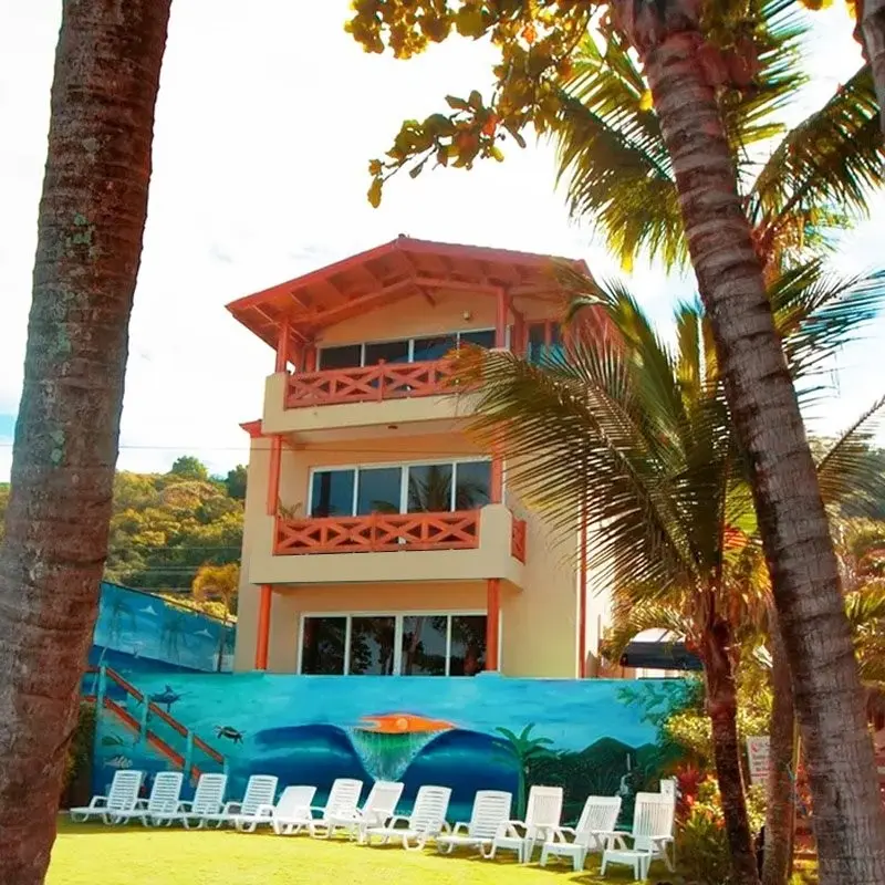 Edificio del Surf Inn Hermosa con un mural de olas, entre palmeras, con sillas blancas sobre el césped frente a la playa