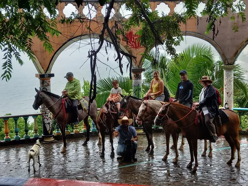 Grupo a caballo bajo un arco de piedra con vista al mar y palmeras, con un perro al frente
