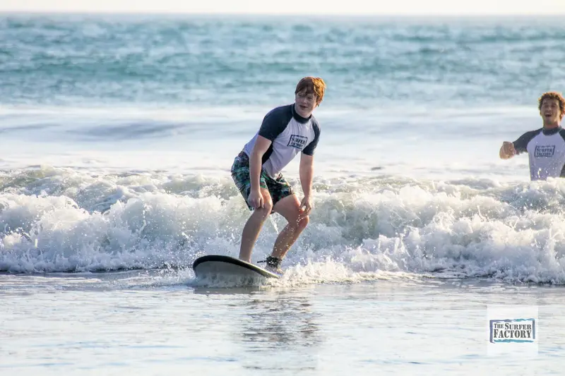 Surfista de pie sobre una tabla en una ola pequeña cerca de la orilla, con otra persona a un lado