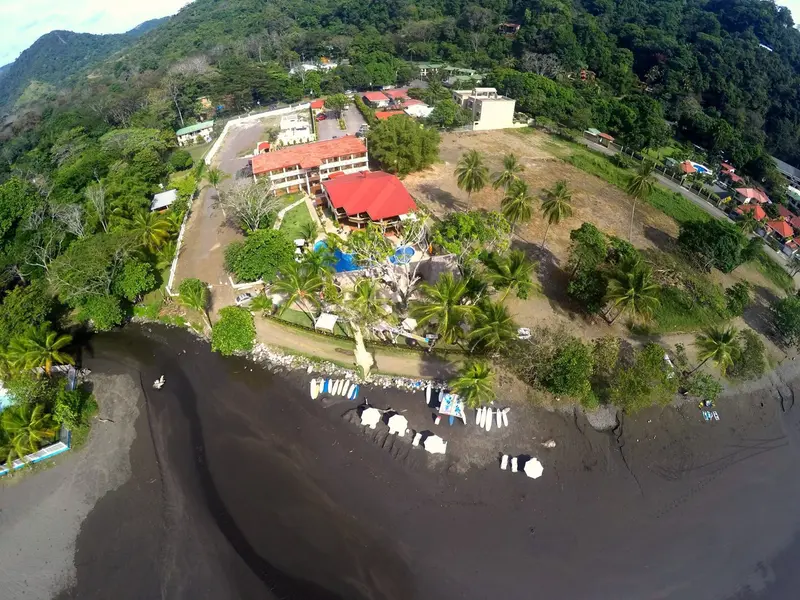 Vista aérea del hotel entre la vegetación, con la playa de arena oscura y sombrillas en la orilla